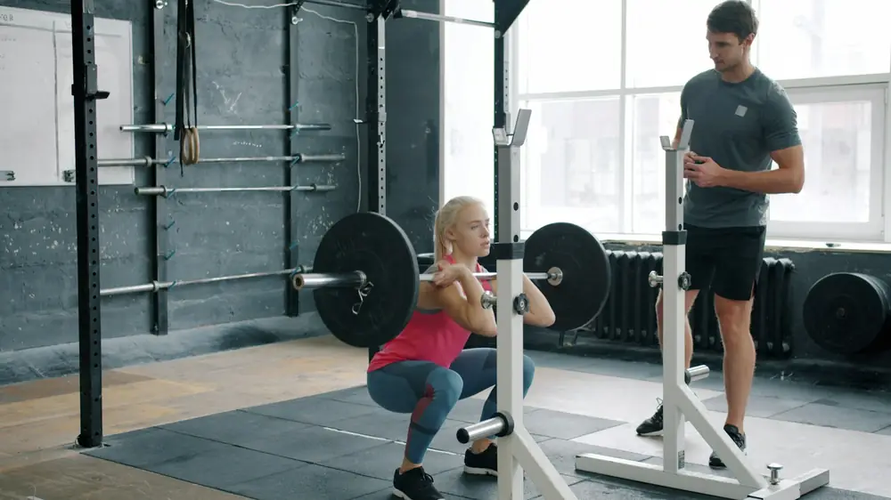 Woman performing barbell squat with trainer watching